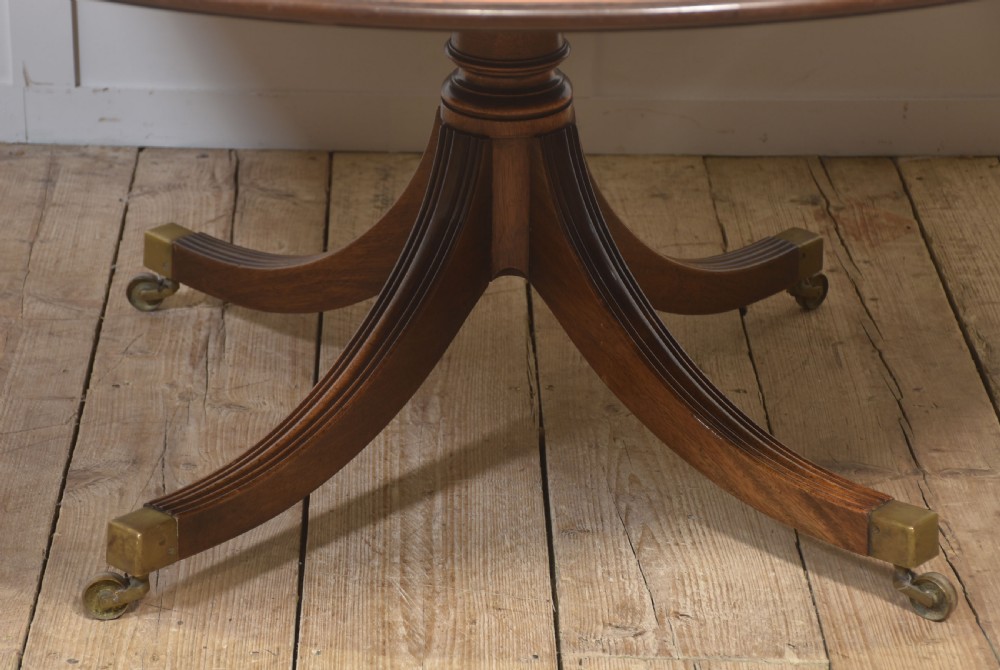 A circular Georgian-style mahogany dining table featuring a crossbanded inlay border, supported by a turned central pedestal with four reeded splayed legs on brass castors. View VI