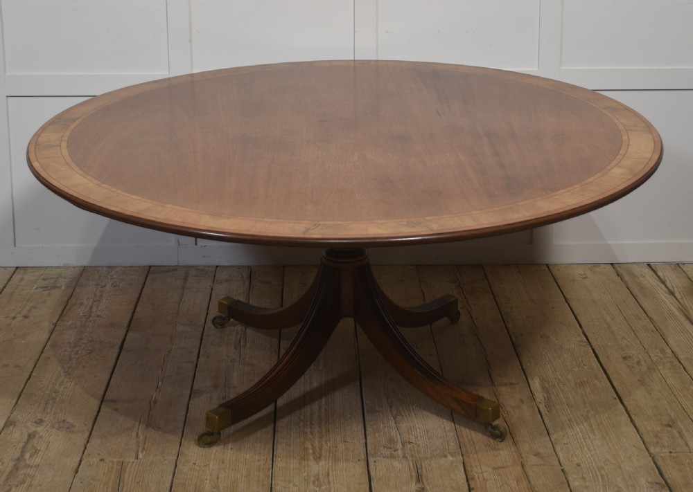 A circular Georgian-style mahogany dining table featuring a crossbanded inlay border, supported by a turned central pedestal with four reeded splayed legs on brass castors. View VII