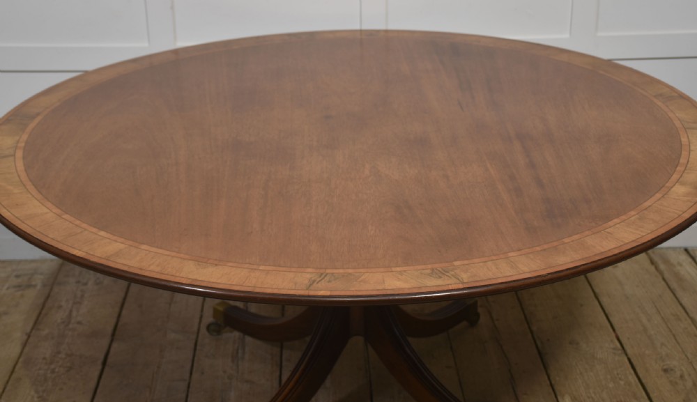 A circular Georgian-style mahogany dining table featuring a crossbanded inlay border, supported by a turned central pedestal with four reeded splayed legs on brass castors. View IX