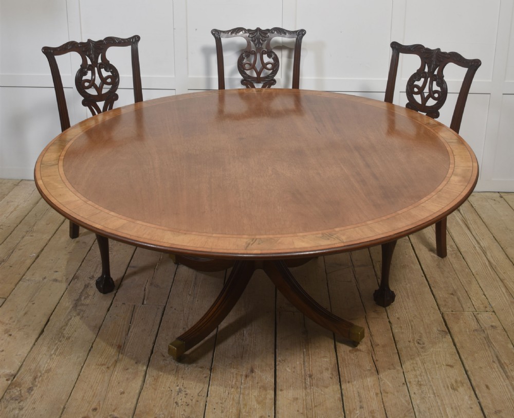 A circular Georgian-style mahogany dining table featuring a crossbanded inlay border, supported by a turned central pedestal with four reeded splayed legs on brass castors. View X