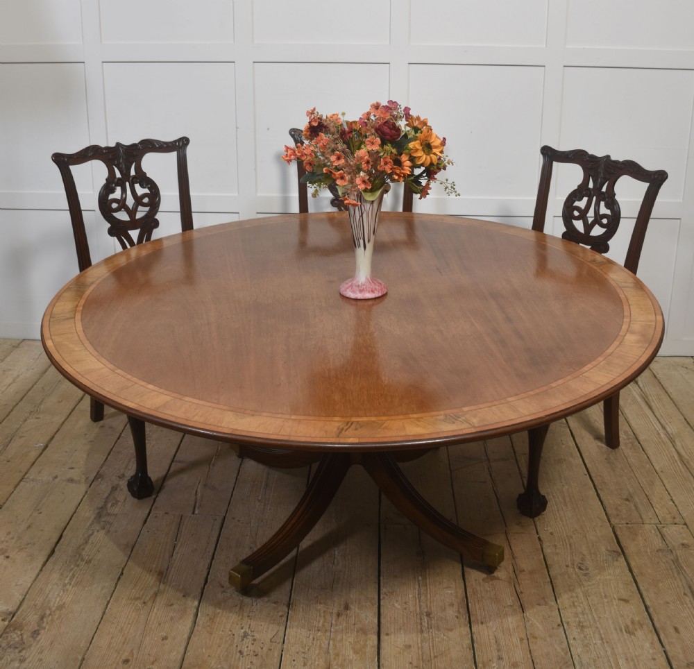 A circular Georgian-style mahogany dining table featuring a crossbanded inlay border, supported by a turned central pedestal with four reeded splayed legs on brass castors. View XI
