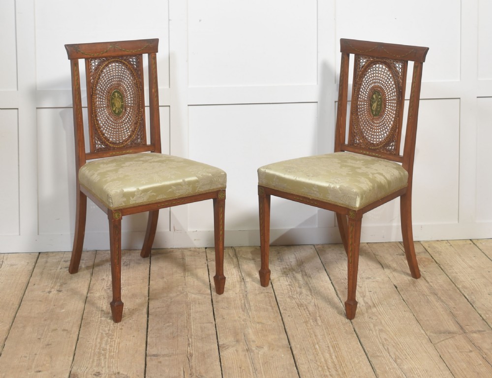 A pair of Edwardian satinwood side chairs featuring oval cane back panels with green painted central medallions, tapered legs, and pale green patterned silk seats.