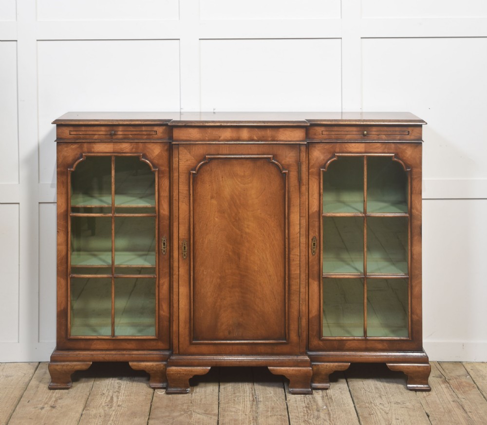 An early 20th-century walnut breakfront cabinet featuring a central solid wooden door flanked by two glazed doors, supported by bracket feet. View V