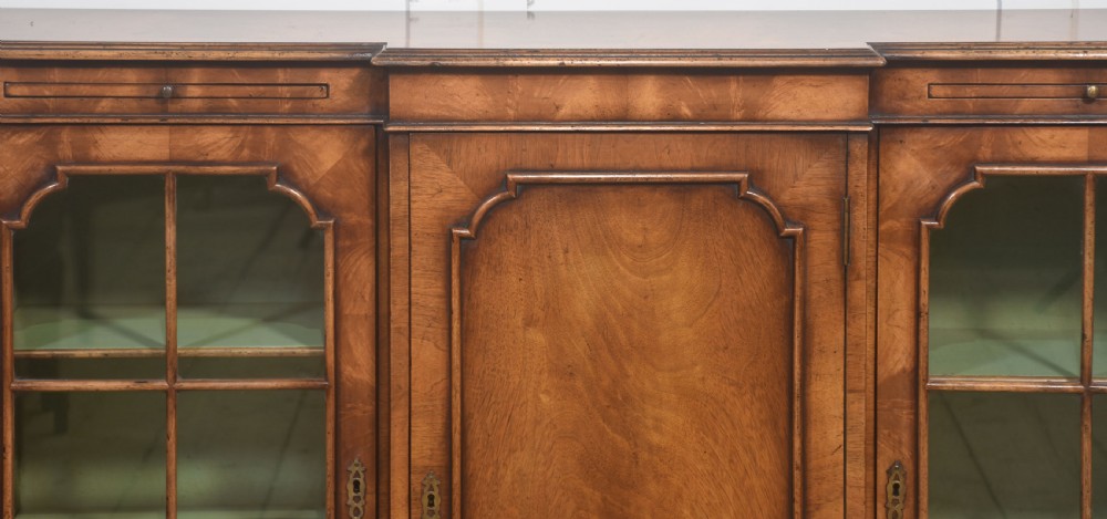 An early 20th-century walnut breakfront cabinet featuring a central solid wooden door flanked by two glazed doors, supported by bracket feet. View VI