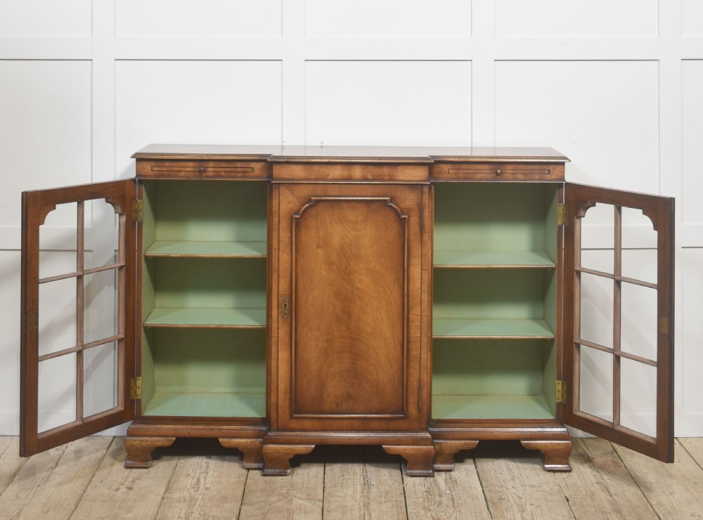 An early 20th-century walnut breakfront cabinet featuring a central solid wooden door flanked by two glazed doors, supported by bracket feet. View IX