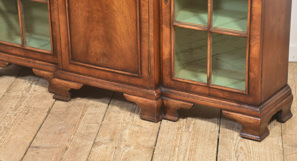 An early 20th-century walnut breakfront cabinet featuring a central solid wooden door flanked by two glazed doors, supported by bracket feet. View XI