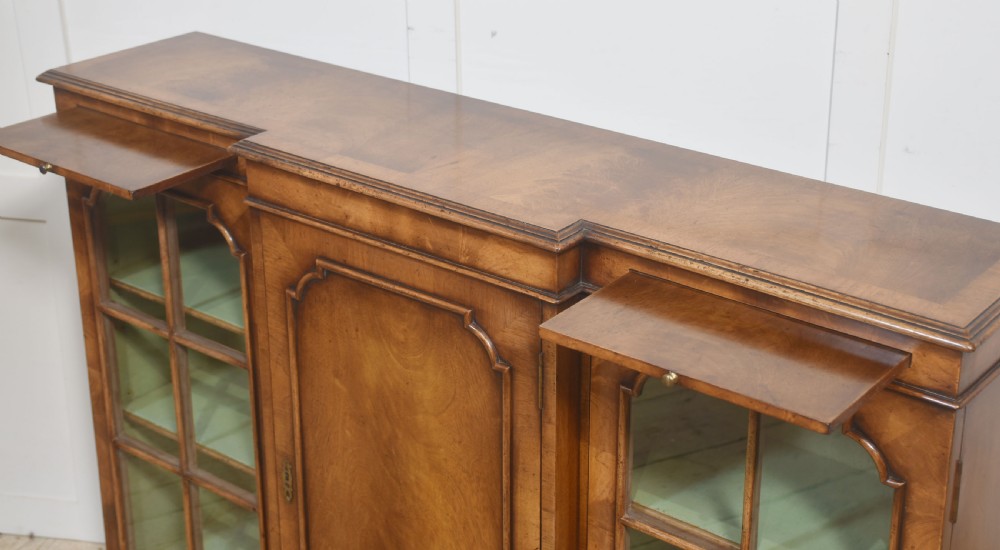 An early 20th-century walnut breakfront cabinet featuring a central solid wooden door flanked by two glazed doors, supported by bracket feet. View XII