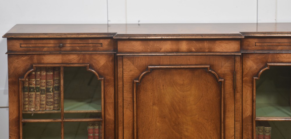 An early 20th-century walnut breakfront cabinet featuring a central solid wooden door flanked by two glazed doors, supported by bracket feet. View XIV