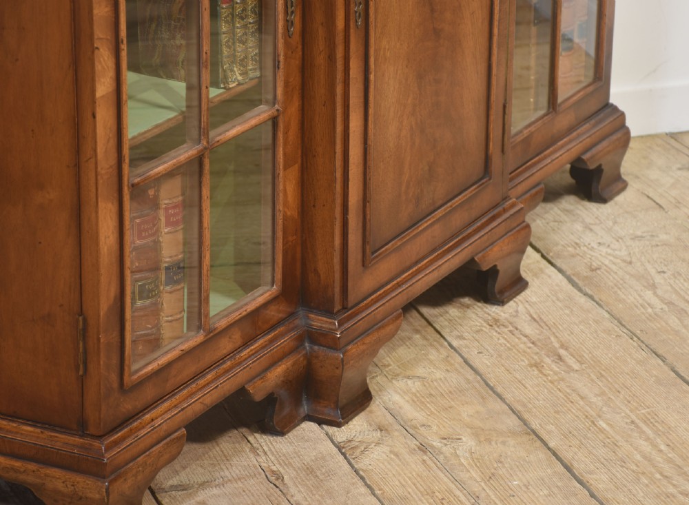 An early 20th-century walnut breakfront cabinet featuring a central solid wooden door flanked by two glazed doors, supported by bracket feet. View XVI