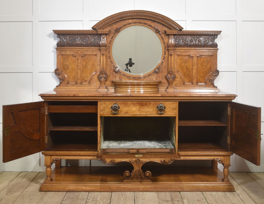 A Victorian carved pollard oak sideboard featuring an upper circular mirror back superstructure, drawers, glazed cabinet doors, and a lower pot board shelf. View VIII