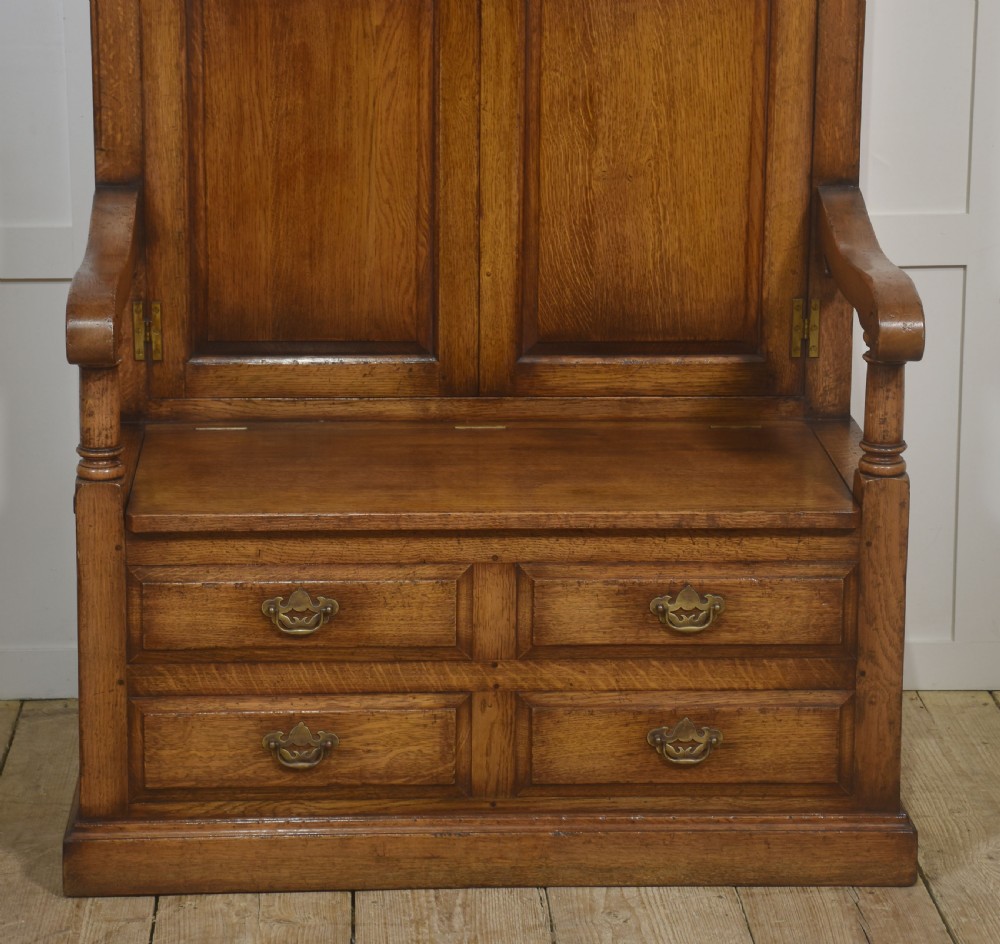 A tall solid oak hall seat cupboard featuring upper twin cupboard doors with exposed brass hinges, a flat wooden bench seat with turned arm supports, and lower drawers with brass drop handles. View IV