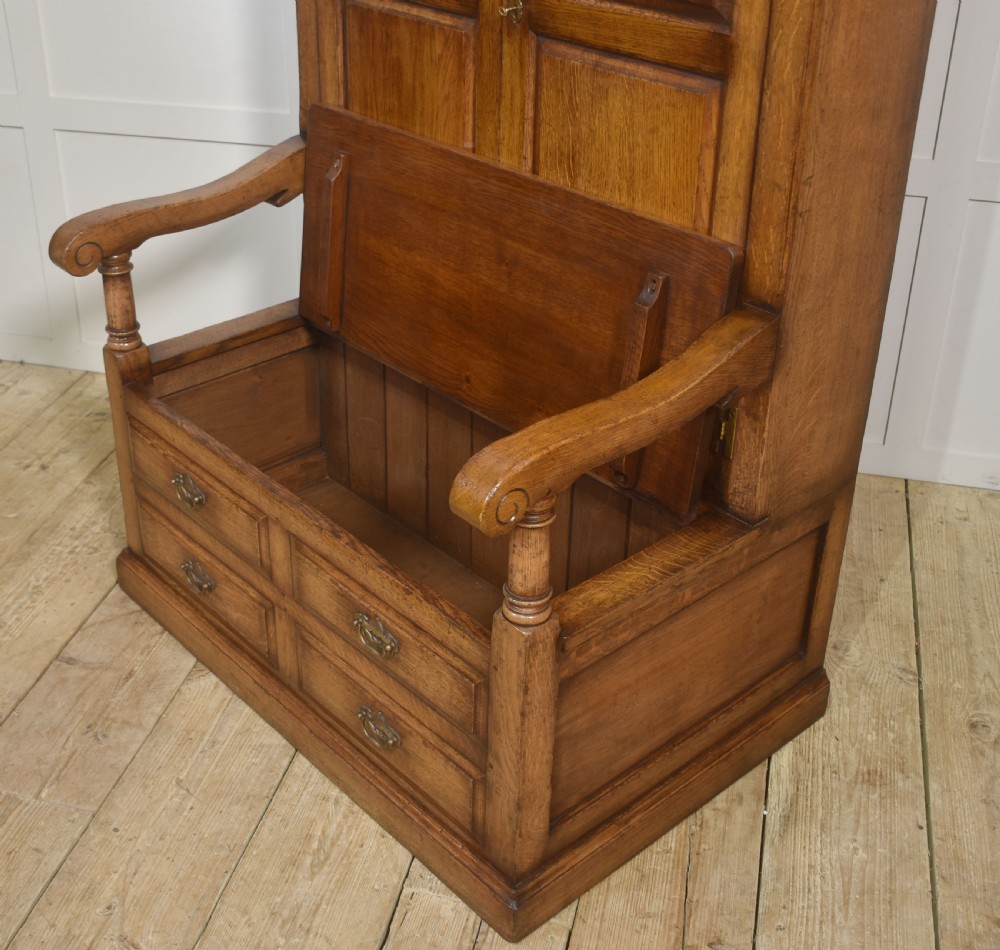 A tall solid oak hall seat cupboard featuring upper twin cupboard doors with exposed brass hinges, a flat wooden bench seat with turned arm supports, and lower drawers with brass drop handles. View XI