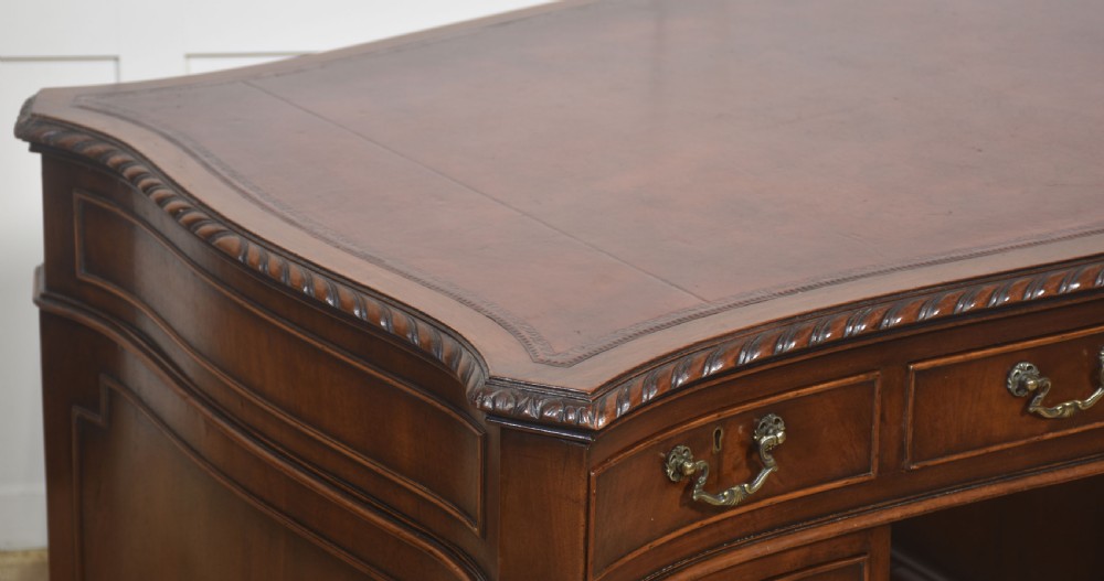 A polished mahogany twin-pedestal desk featuring brass swan-neck handles, carved edge moulding, and multiple drawers set on bracket feet. View VII