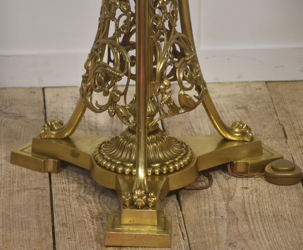 An ornate brass floor lamp featuring a tapered openwork base and a light blue patterned lampshade with fringe detailing. View III