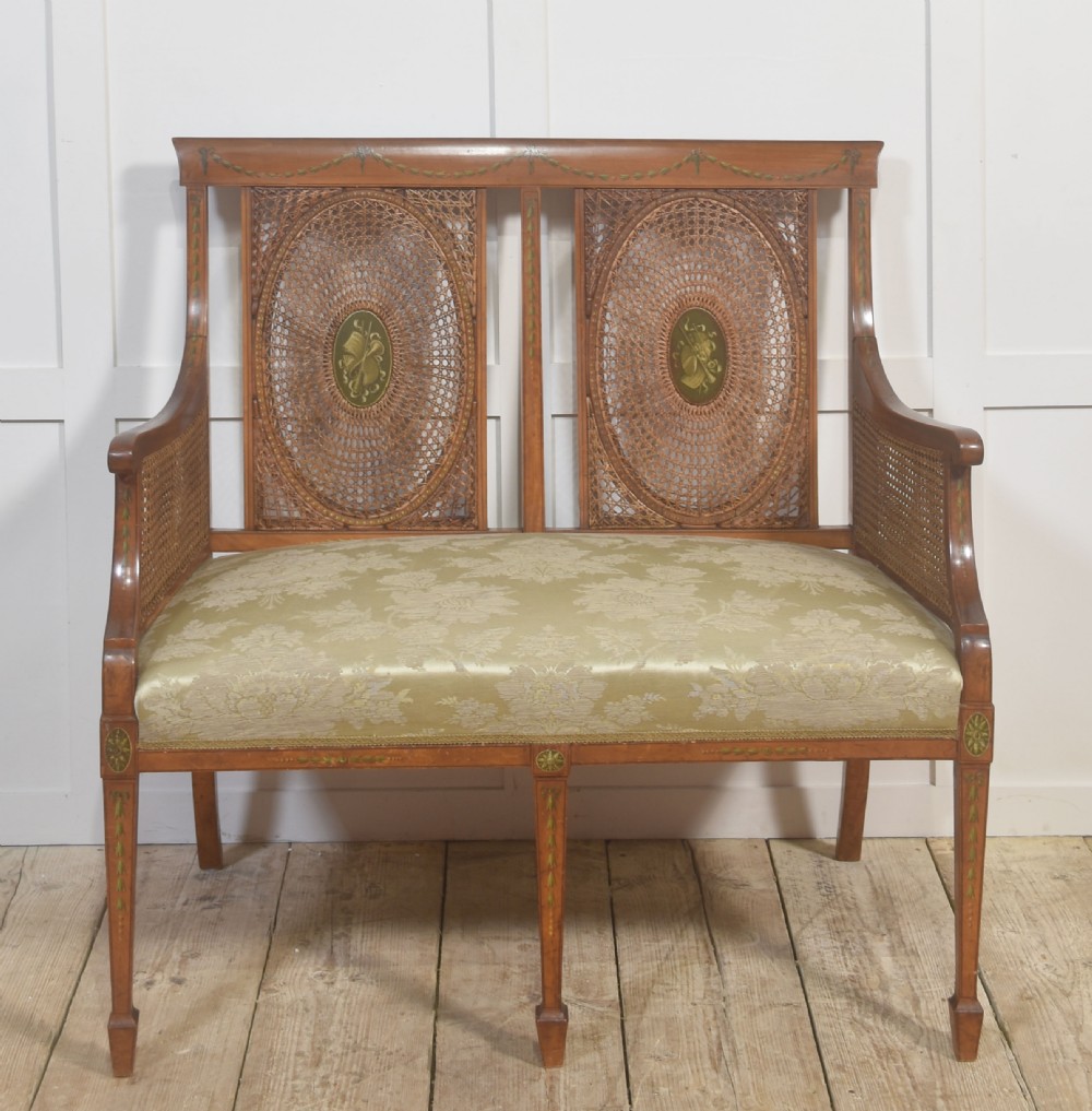 A painted satinwood two-chair-back settee featuring oval caned panels, hand-painted green floral garlands, swept armrests, and an upholstered damask seat.