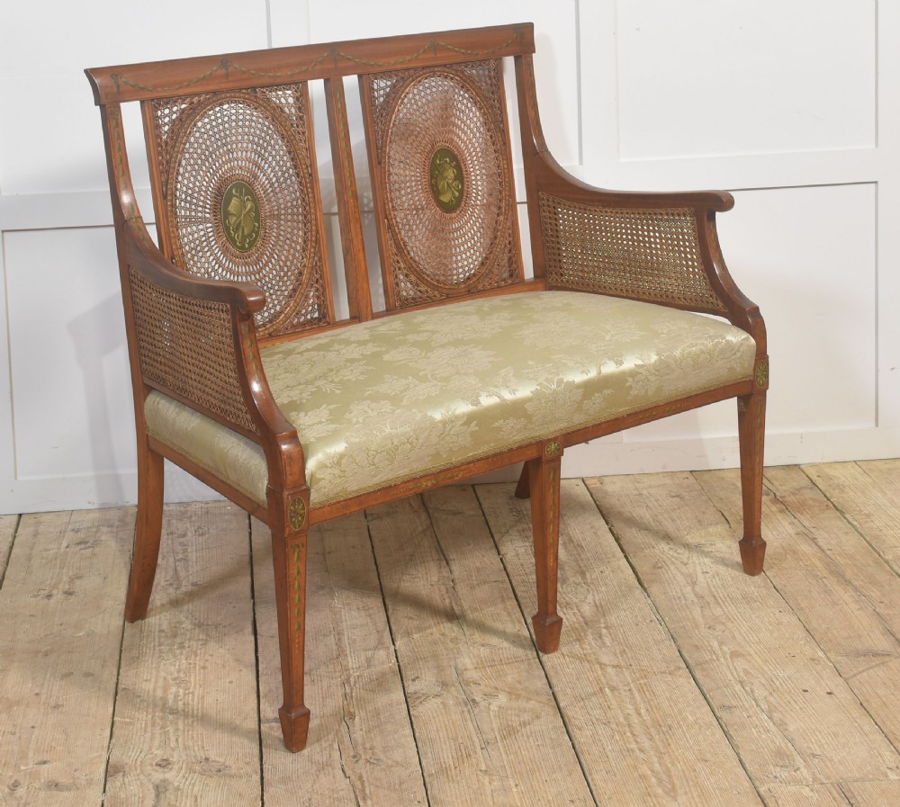 A painted satinwood two-chair-back settee featuring oval caned panels, hand-painted green floral garlands, swept armrests, and an upholstered damask seat. View III
