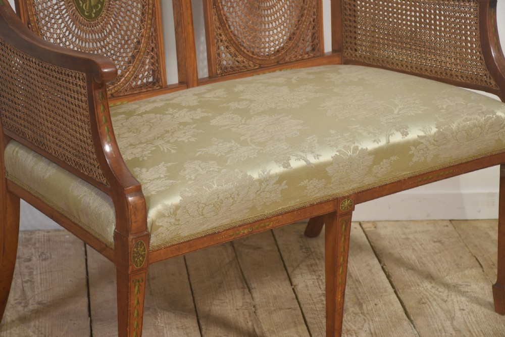 A painted satinwood two-chair-back settee featuring oval caned panels, hand-painted green floral garlands, swept armrests, and an upholstered damask seat. View IV