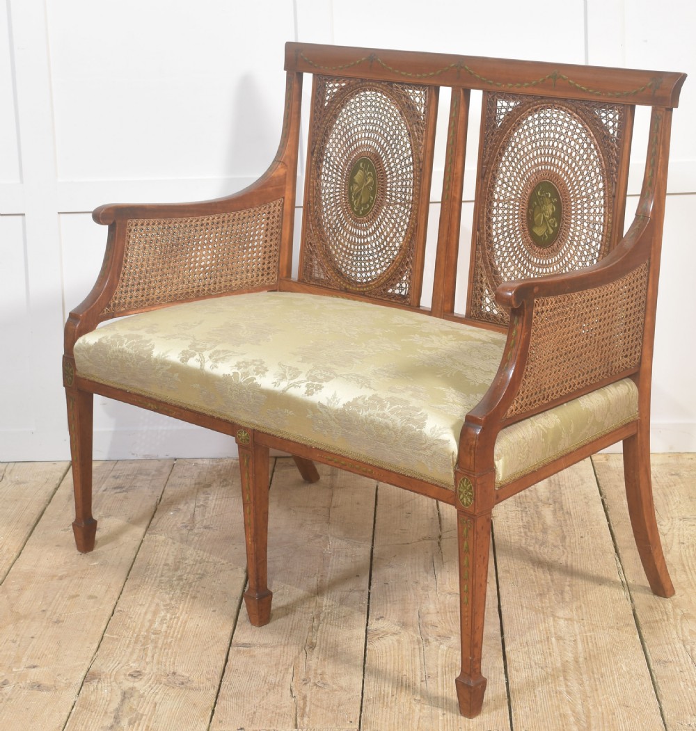 A painted satinwood two-chair-back settee featuring oval caned panels, hand-painted green floral garlands, swept armrests, and an upholstered damask seat. View VI