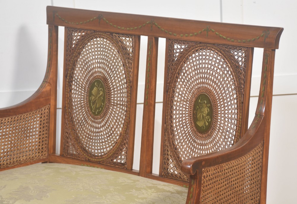 A painted satinwood two-chair-back settee featuring oval caned panels, hand-painted green floral garlands, swept armrests, and an upholstered damask seat. View VII