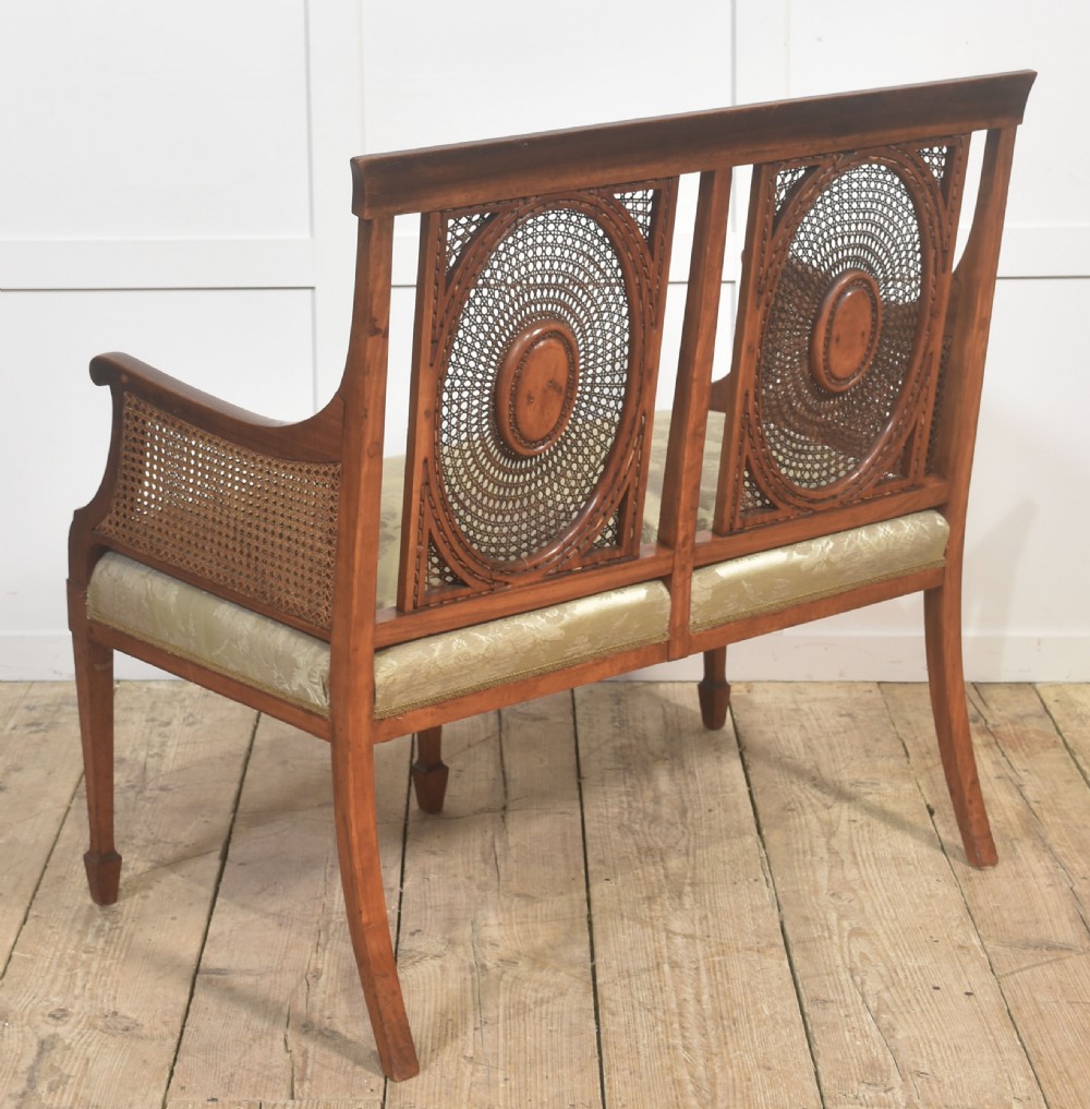 A painted satinwood two-chair-back settee featuring oval caned panels, hand-painted green floral garlands, swept armrests, and an upholstered damask seat. View VIII