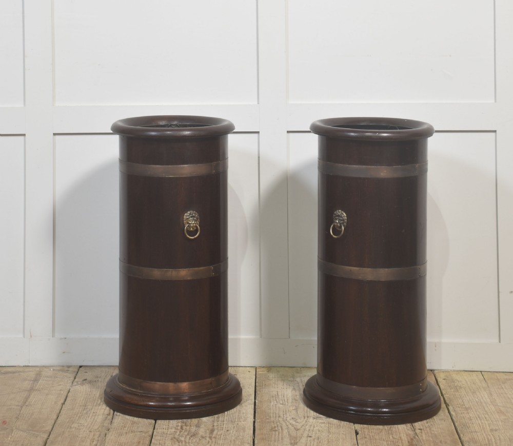A pair of cylindrical dark mahogany umbrella stands featuring brass binding bands and brass lion mask ring handles.