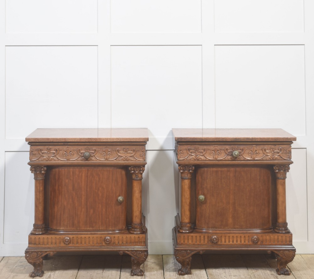 A pair of carved warm brown mahogany bedside cabinets featuring classical columns, frieze carvings, and single panel doors on carved feet.