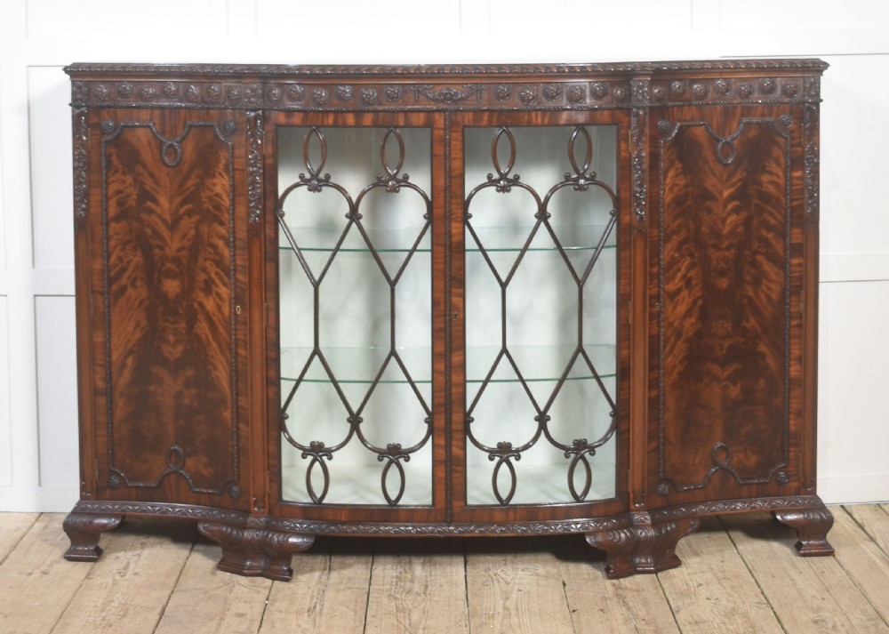 A wide mahogany display cabinet featuring flame mahogany veneered side doors, central astragal glazed double doors, carved rosettes along the frieze, and cabriole feet.