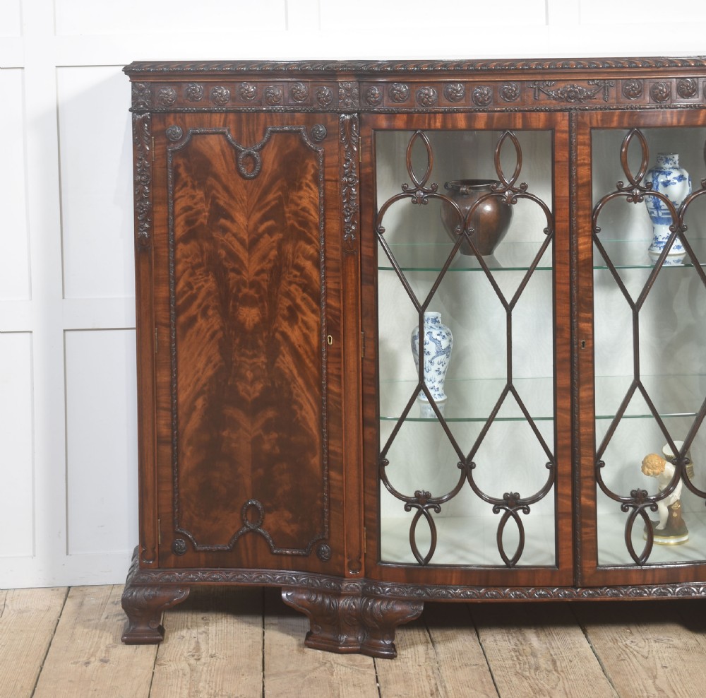 A wide mahogany display cabinet featuring flame mahogany veneered side doors, central astragal glazed double doors, carved rosettes along the frieze, and cabriole feet. View IV