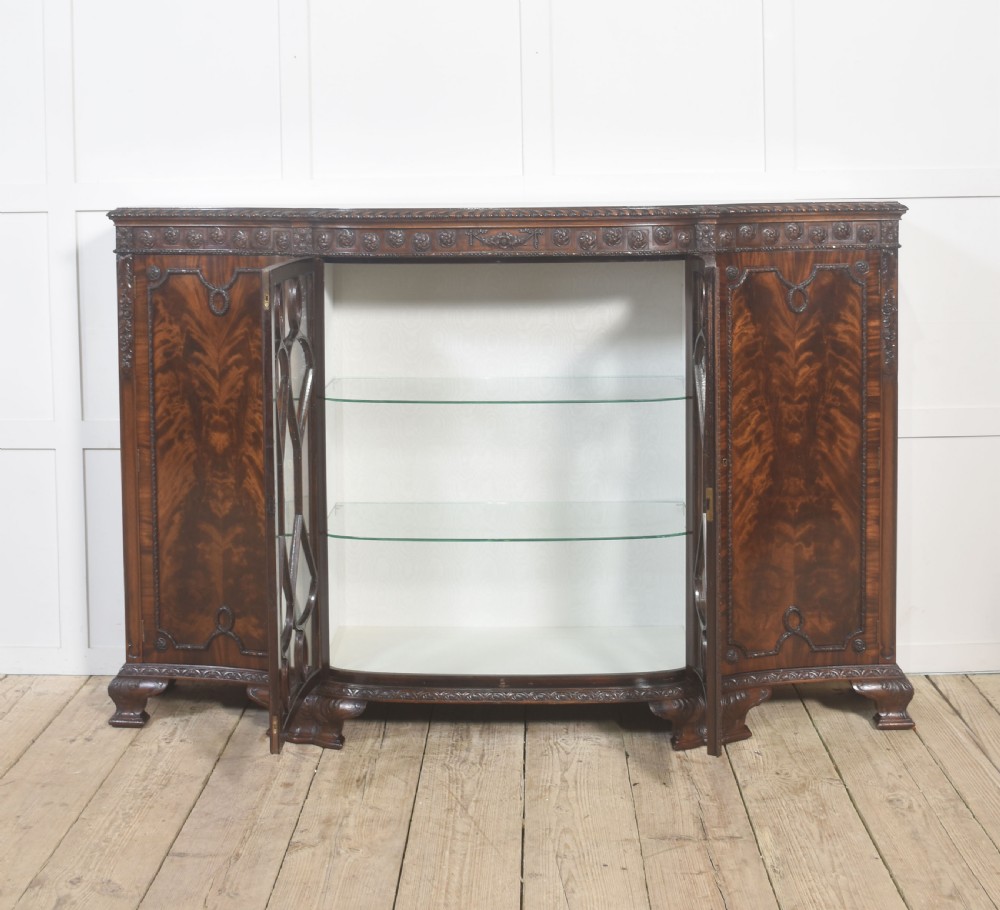 A wide mahogany display cabinet featuring flame mahogany veneered side doors, central astragal glazed double doors, carved rosettes along the frieze, and cabriole feet. View VII