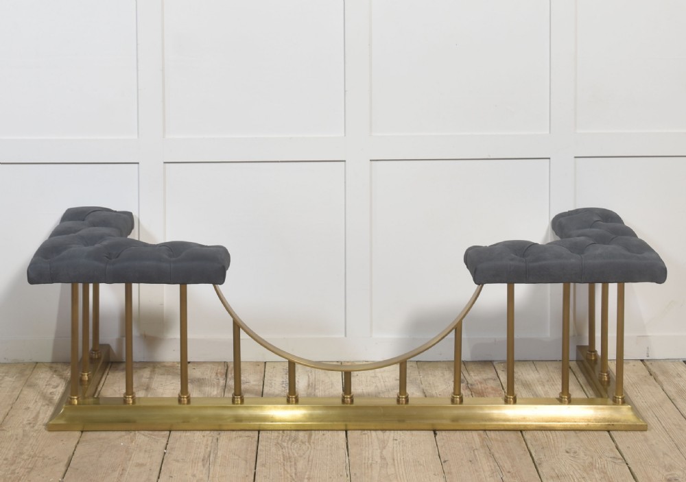 A solid brass club fender featuring twin button-upholstered grey corner seats supported by vertical brass spindles and a dipped central railing.