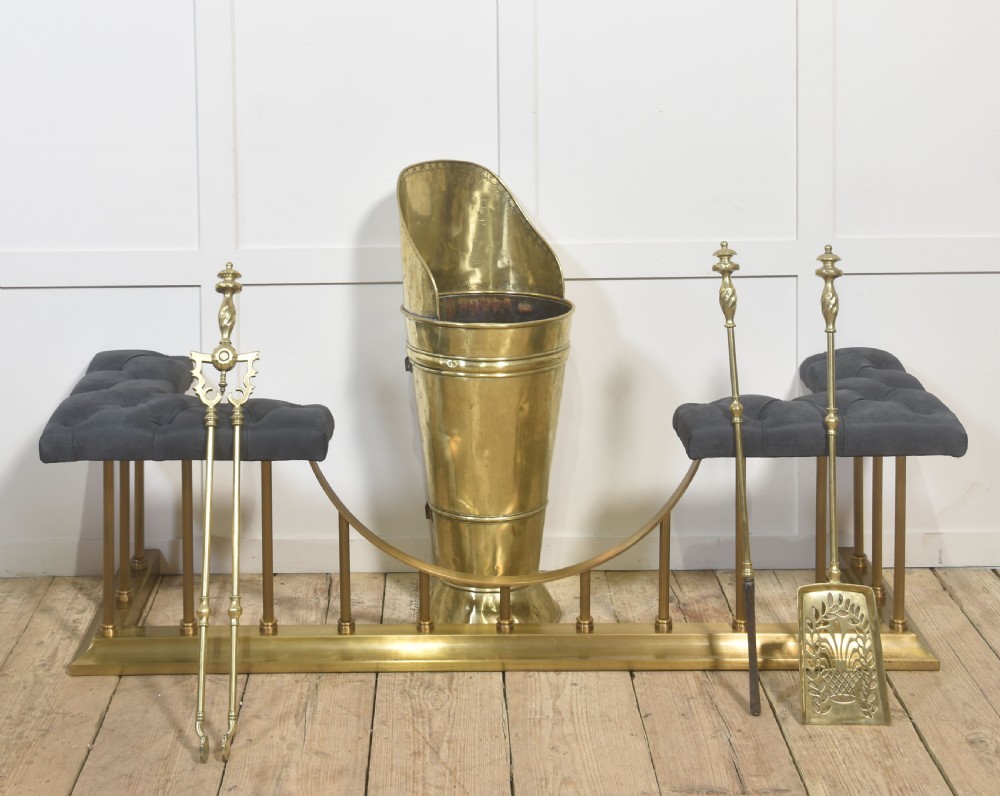 A solid brass club fender featuring twin button-upholstered grey corner seats supported by vertical brass spindles and a dipped central railing. View II