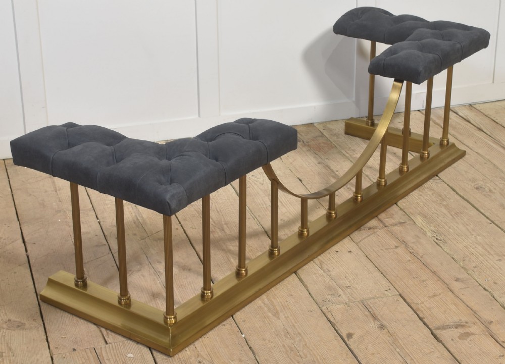 A solid brass club fender featuring twin button-upholstered grey corner seats supported by vertical brass spindles and a dipped central railing. View V