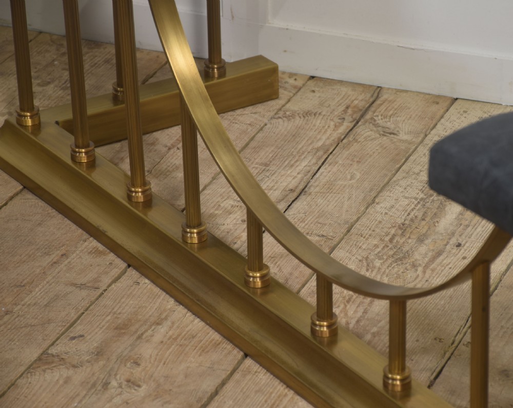 A solid brass club fender featuring twin button-upholstered grey corner seats supported by vertical brass spindles and a dipped central railing. View X