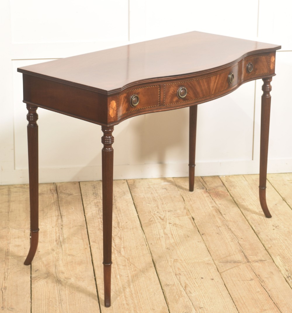 An inlaid mahogany writing table featuring a serpentine front, drawers with brass ring handles, oval patera marquetry, and fluted legs. View IV