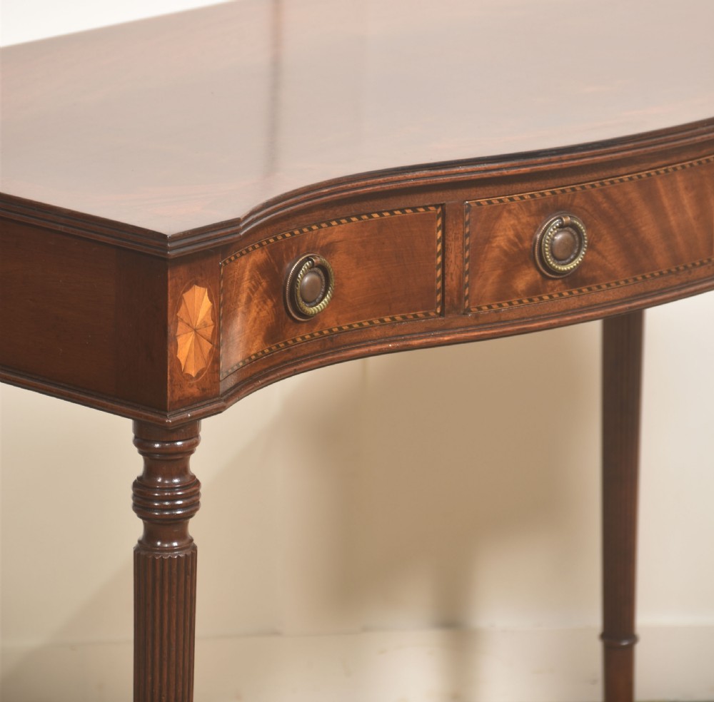 An inlaid mahogany writing table featuring a serpentine front, drawers with brass ring handles, oval patera marquetry, and fluted legs. View VII