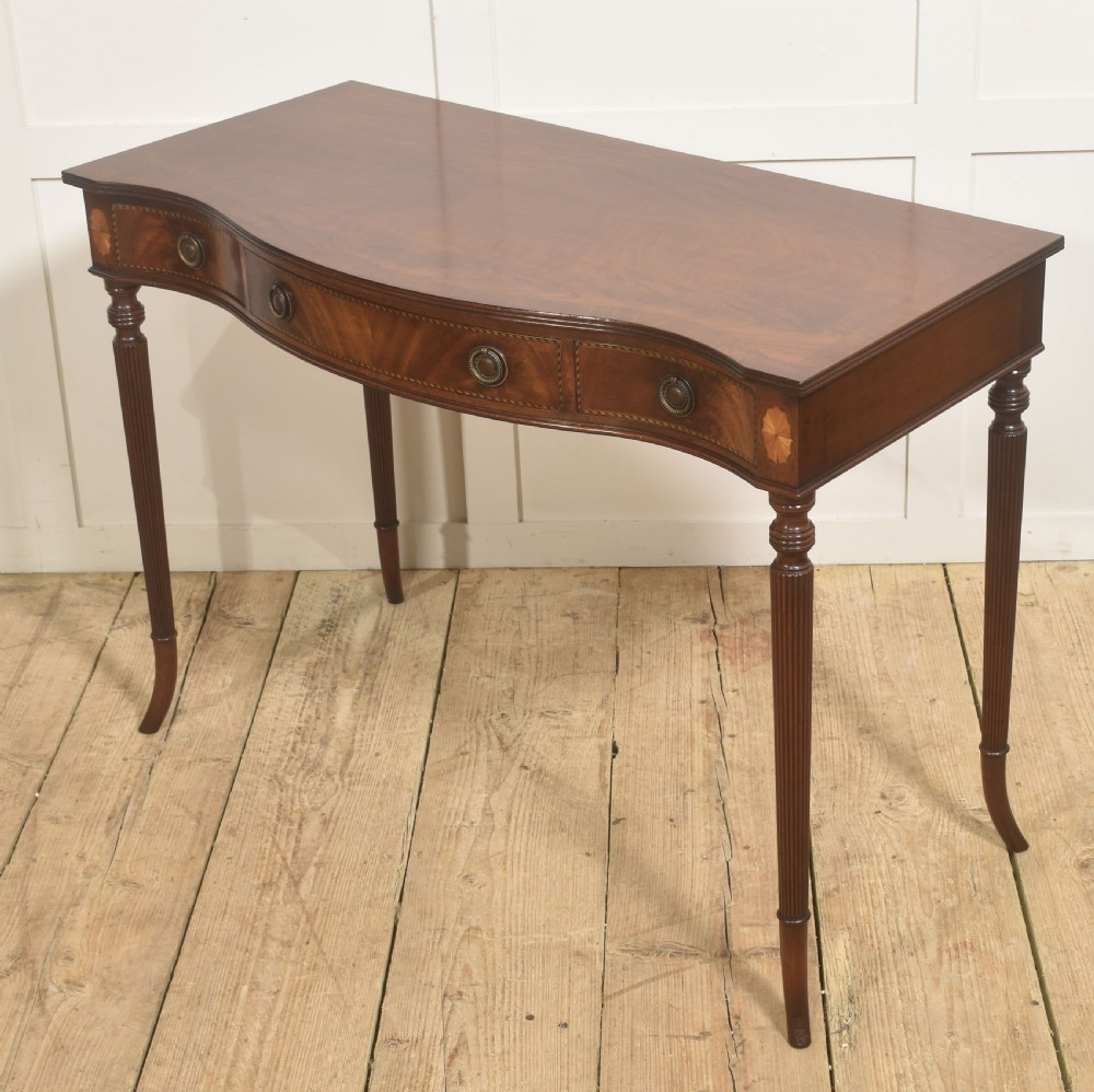 An inlaid mahogany writing table featuring a serpentine front, drawers with brass ring handles, oval patera marquetry, and fluted legs. View IX
