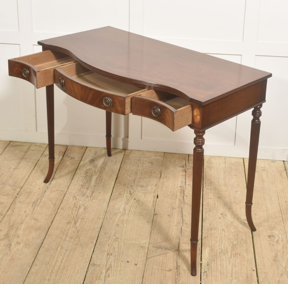 An inlaid mahogany writing table featuring a serpentine front, drawers with brass ring handles, oval patera marquetry, and fluted legs. View X