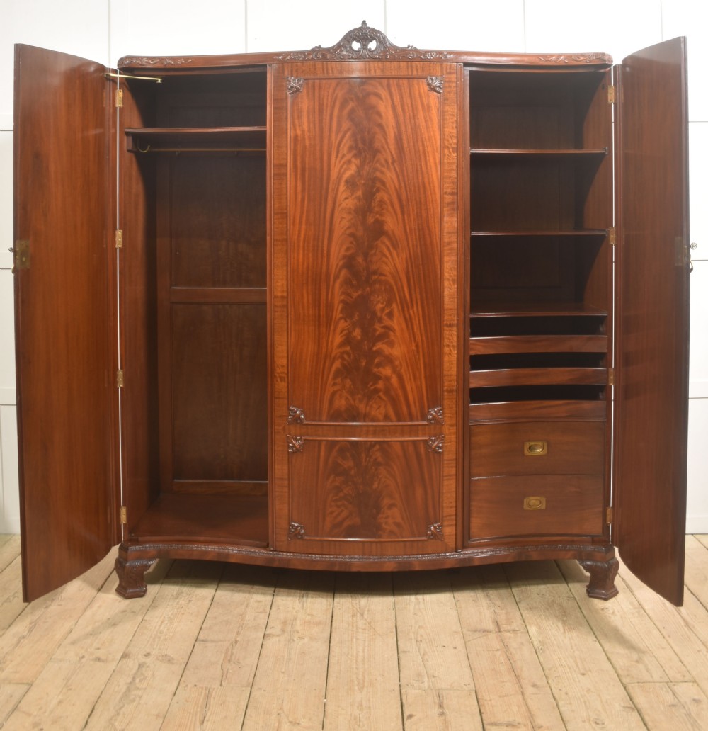 An antique flame mahogany three-door wardrobe featuring a carved central crest, moulded panel doors, and brass drop handles. View IV