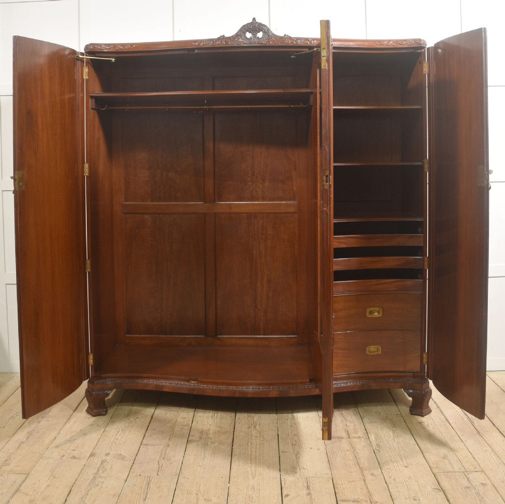 An antique flame mahogany three-door wardrobe featuring a carved central crest, moulded panel doors, and brass drop handles. View V