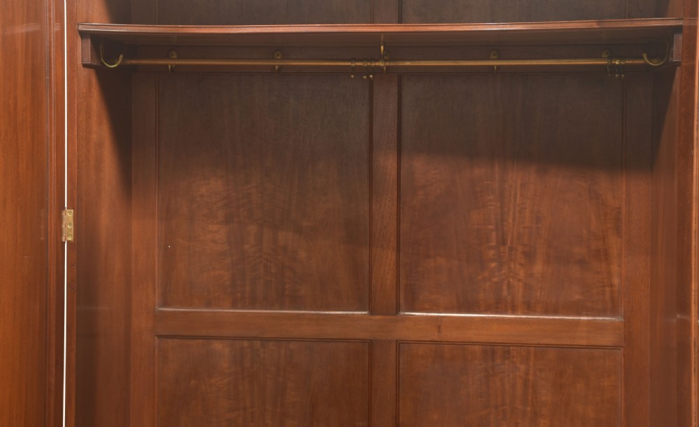 An antique flame mahogany three-door wardrobe featuring a carved central crest, moulded panel doors, and brass drop handles. View VI