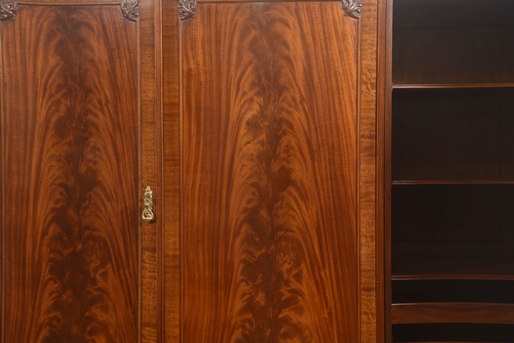 An antique flame mahogany three-door wardrobe featuring a carved central crest, moulded panel doors, and brass drop handles. View VII