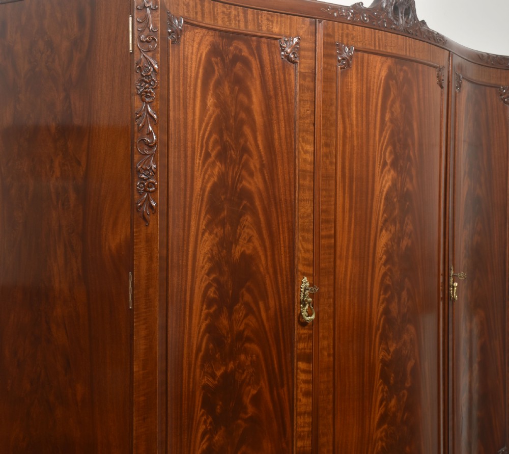 An antique flame mahogany three-door wardrobe featuring a carved central crest, moulded panel doors, and brass drop handles. View X
