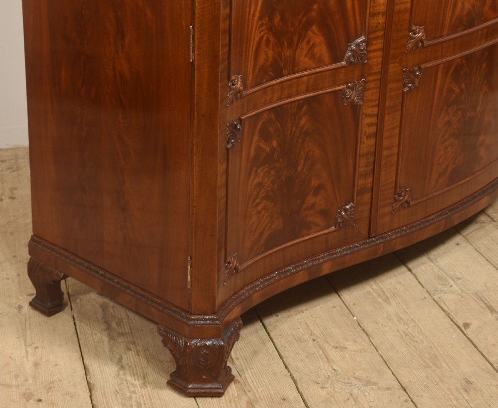 An antique flame mahogany three-door wardrobe featuring a carved central crest, moulded panel doors, and brass drop handles. View XII