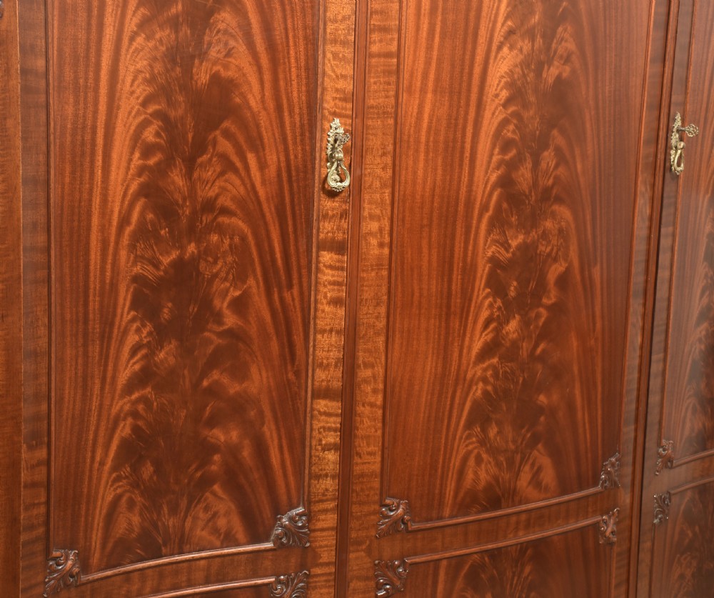 An antique flame mahogany three-door wardrobe featuring a carved central crest, moulded panel doors, and brass drop handles. View XIII