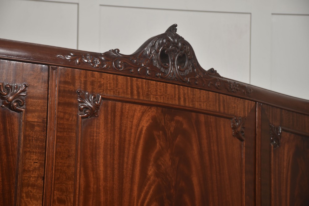 An antique flame mahogany three-door wardrobe featuring a carved central crest, moulded panel doors, and brass drop handles. View XIV