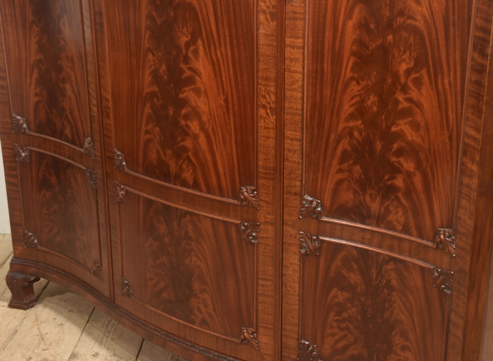 An antique flame mahogany three-door wardrobe featuring a carved central crest, moulded panel doors, and brass drop handles. View XVI