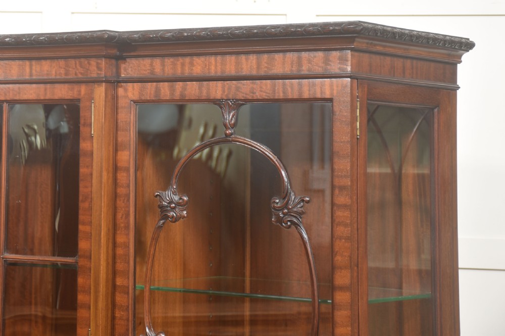 A Chippendale Revival breakfront display cabinet in carved mahogany featuring glazed doors, interior glass shelves, lower cupboards, and claw-and-ball feet. View IX
