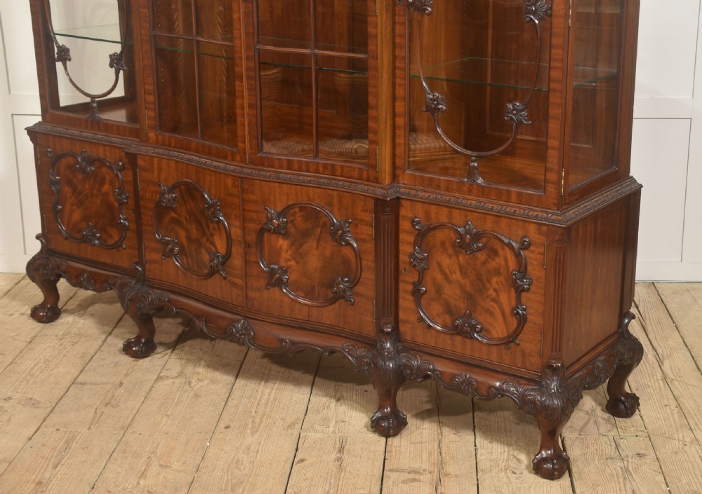 A Chippendale Revival breakfront display cabinet in carved mahogany featuring glazed doors, interior glass shelves, lower cupboards, and claw-and-ball feet. View X