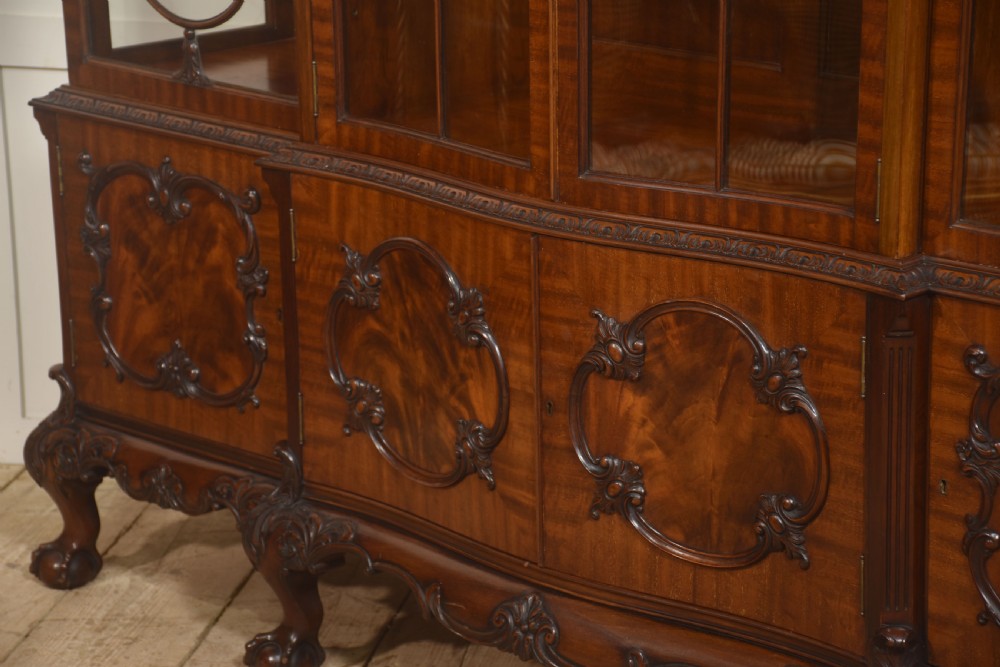 A Chippendale Revival breakfront display cabinet in carved mahogany featuring glazed doors, interior glass shelves, lower cupboards, and claw-and-ball feet. View XIII