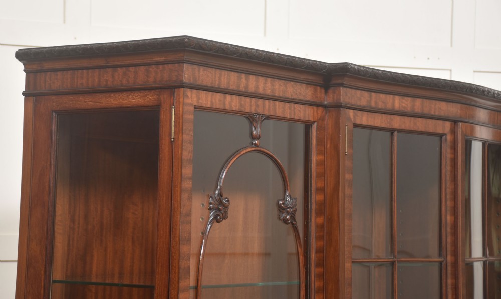 A Chippendale Revival breakfront display cabinet in carved mahogany featuring glazed doors, interior glass shelves, lower cupboards, and claw-and-ball feet. View XV